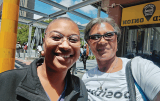 Two women smiling for a selfie outdoors in an urban plaza, wearing glasses and a necklace, with a yellow pole and storefront in the background.
