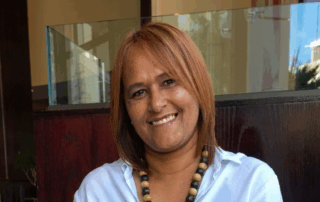 Smiling woman with shoulder-length light brown hair, wearing a light blue shirt and a beaded necklace.