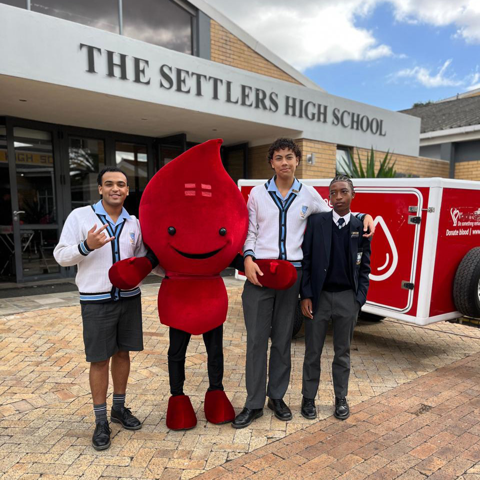 settlers-high-donates-blood Three students in school uniforms pose with a red blood-drop mascot outside a high school, beside a blood donation trailer.